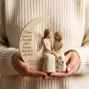May include: A cream-colored figurine of two figures sitting on a crescent moon. One figure has wings. The moon is engraved with the text: I know heaven is a beautiful place because they have my mom, Anna Scott, 1954-2026. The second figure features floral details.