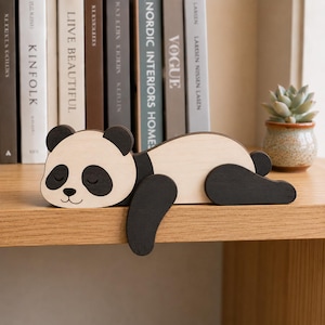 Op de afbeelding: Een plat houten panda-figuurtje met een lichtbeige lichaam en donkerbruine oren, oogvlekken, ledematen en een kleine neus. De panda is afgebeeld in een slaaphouding met de voorpoten over de rand van een plat oppervlak hangend.
