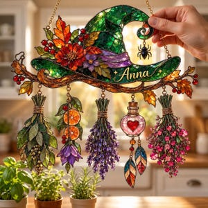 Puede incluir: Un atrapasoles de vidrieras con un sombrero de bruja verde adornado con hojas de otoño, bayas y el nombre Anna. Debajo cuelgan cinco elementos decorativos: manojos de hierbas, rodajas de naranja, una flor morada, un frasco y plumas.