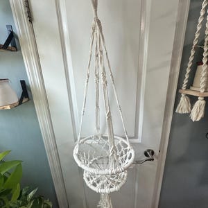 May include: A white macrame plant hanger featuring a two-tiered circular design with knotted cord construction. The hanger includes a top loop for suspension, four main support cords, and a decorative tassel at the bottom center.