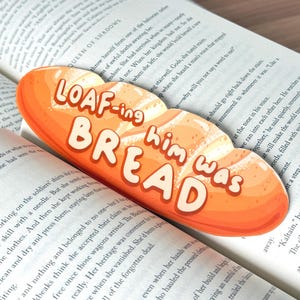 May include: A whimsical bookmark shaped like a loaf of bread, with the words "LOAF-ing him was BREAD" in playful lettering. The bookmark is orange and white, resting on the pages of an open book.