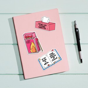 May include: Four pink and white stickers with book-themed sayings. The stickers feature a book, a box of matches, a tissue box, and a box of matches. The text on the stickers reads: "If my book is open, your mouth should be closed", "Slow Burn", "Crying over fictional characters", and "Glow Burn".
