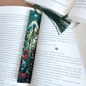 May include: A decorative bookmark featuring a detailed illustration of a woman with a sword, surrounded by stained glass-style elements and red roses. A dark green tassel hangs from the top. The bookmark is placed in an open book.