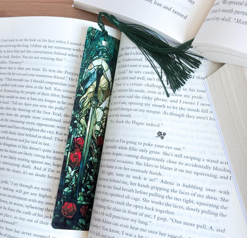 May include: A decorative bookmark featuring a detailed illustration of a woman with a sword, surrounded by stained glass-style elements and red roses. A dark green tassel hangs from the top. The bookmark is placed in an open book.