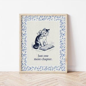 May include: Framed print featuring a black and white illustration of a cat with a bow reading a book. The print has a floral border with blue flowers and the text "Just one more chapter."