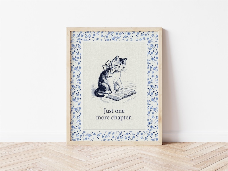 May include: Framed print featuring a black and white illustration of a cat with a bow reading a book. The print has a floral border with blue flowers and the text "Just one more chapter."