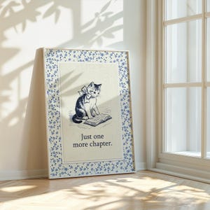 May include: Framed print featuring a black and white illustration of a cat with a bow, sitting on a book. The frame has a blue floral pattern. Text reads: "Just one more chapter."