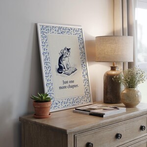 May include: A framed print featuring a black and white illustration of a cat reading a book, with the text "Just one more chapter." The frame has a white border with a blue floral pattern. The print is displayed on a wooden dresser.