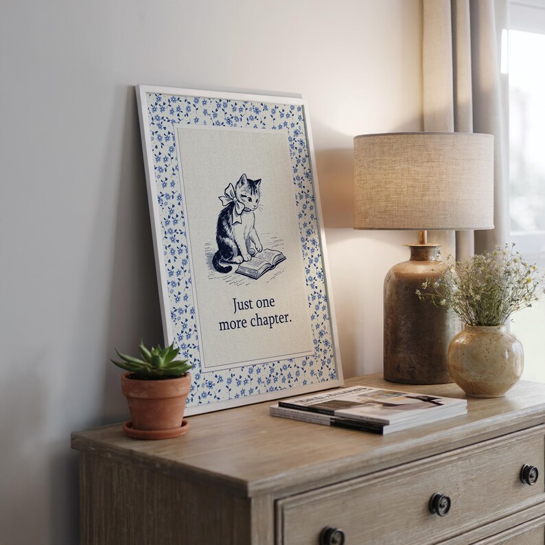 May include: A framed print featuring a black and white illustration of a cat reading a book, with the text "Just one more chapter." The frame has a white border with a blue floral pattern. The print is displayed on a wooden dresser.