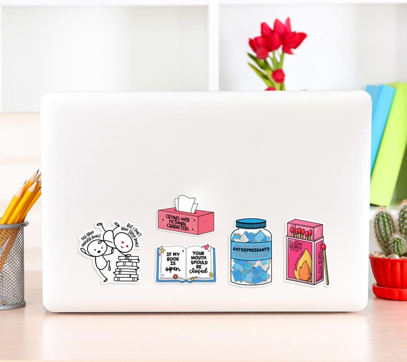 May include: A white laptop with six colorful stickers. The stickers feature illustrations and text. One sticker says "If my book is open, your mouth should be closed". Another sticker says "Crying over fictional characters".