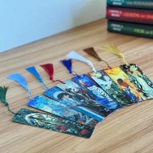 May include: A collection of colorful, illustrated bookmarks with decorative tassels. The bookmarks feature various artistic designs and are fanned out on a wooden surface. Several books are stacked in the background.