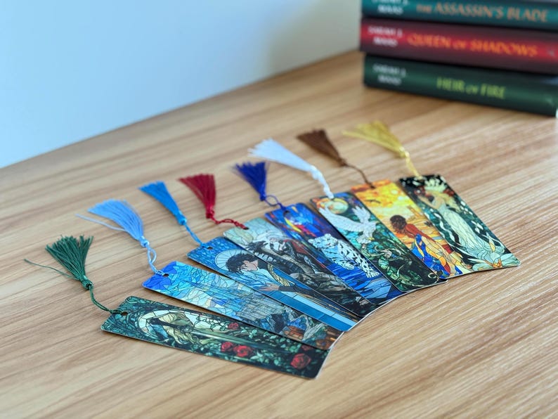 May include: A collection of colorful, illustrated bookmarks with decorative tassels. The bookmarks feature various artistic designs and are fanned out on a wooden surface. Several books are stacked in the background.