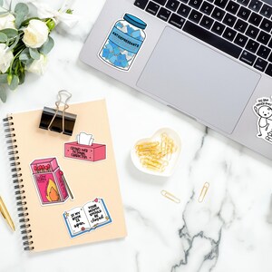May include: A light brown notebook with a spiral binding and a variety of colorful stickers on the cover. The stickers feature phrases like "Crying over fictional characters" and "Your mouth should be closed".