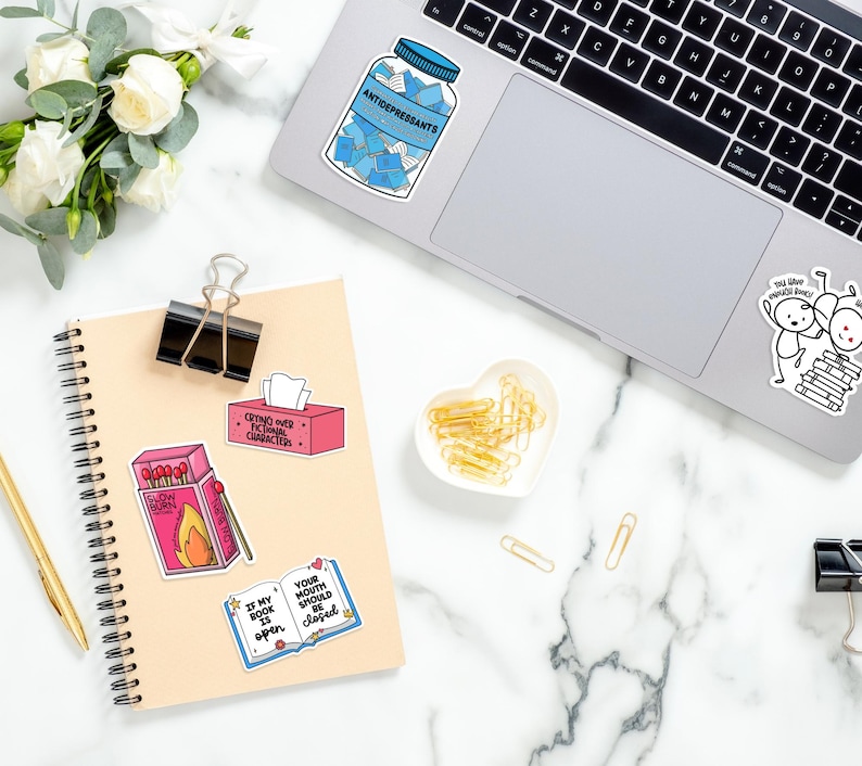 May include: A light brown notebook with a spiral binding and a variety of colorful stickers on the cover. The stickers feature phrases like "Crying over fictional characters" and "Your mouth should be closed".