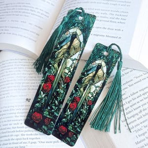 May include: Two decorative bookmarks with a stained-glass design featuring a woman, a sword, and red roses. The bookmarks have green tassels and are set against the backdrop of an open book.