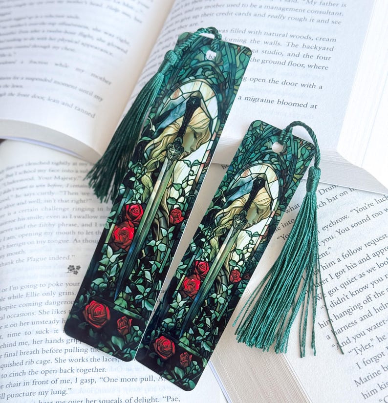 May include: Two decorative bookmarks with a stained-glass design featuring a woman, a sword, and red roses. The bookmarks have green tassels and are set against the backdrop of an open book.