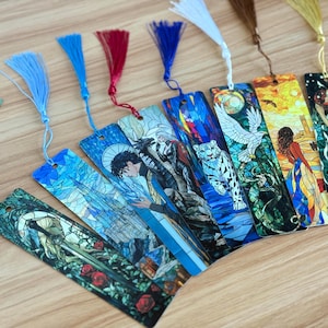 May include: A collection of fantasy-themed bookmarks with colorful tassel accents. Each bookmark features a unique illustration, including a castle, a person, a tiger, and a woman. The bookmarks are arranged on a light-colored wooden surface.