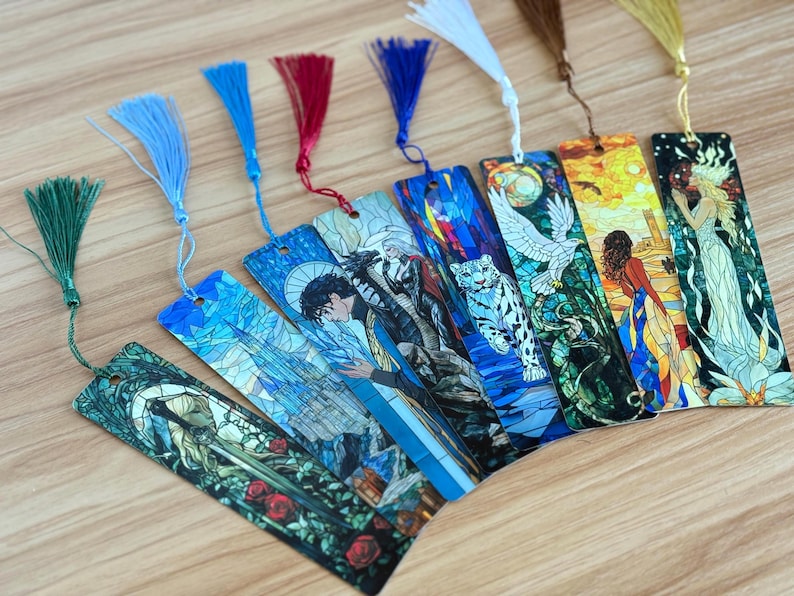 May include: A collection of fantasy-themed bookmarks with colorful tassel accents. Each bookmark features a unique illustration, including a castle, a person, a tiger, and a woman. The bookmarks are arranged on a light-colored wooden surface.