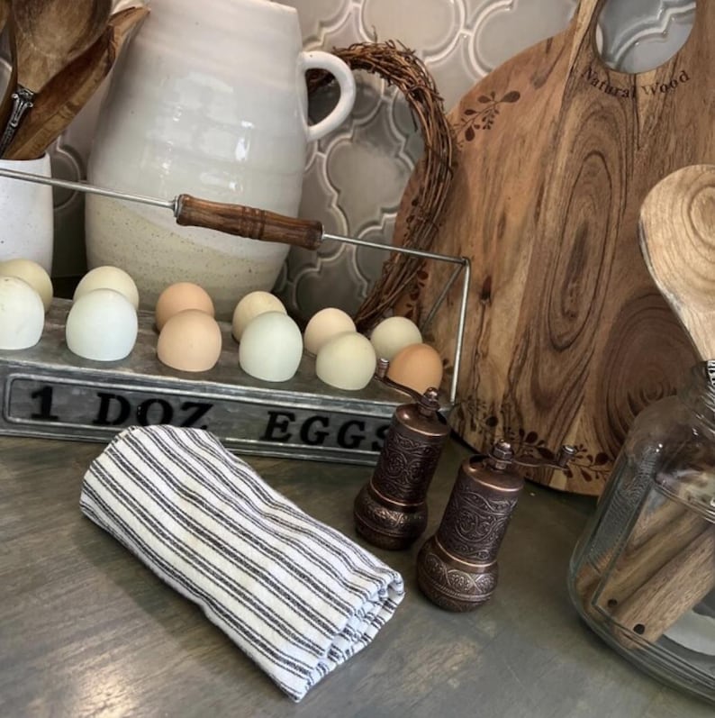 May include: A rectangular metal egg holder with the text 1 DOZ EGGS containing twelve eggs in various shades of white and brown. Beside it are two ornate copper-colored spice grinders and a folded white cloth with thin black vertical stripes.