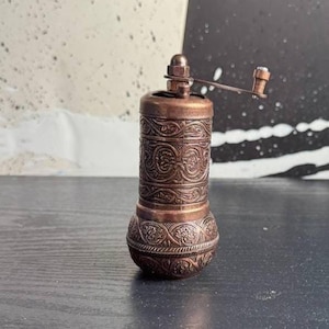 May include: A cylindrical copper-toned spice grinder featuring intricate embossed floral and scrollwork patterns across its body. The manual mill includes a small metal handle at the top with a knob, designed for grinding spices with a rotating motion.