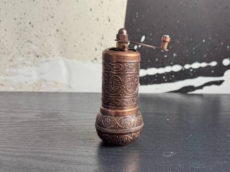May include: A cylindrical copper-toned spice grinder featuring intricate embossed floral and scrollwork patterns across its body. The manual mill includes a small metal handle at the top with a knob, designed for grinding spices with a rotating motion.