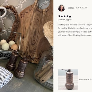May include: Two cylindrical copper spice grinders with intricate embossed patterns, featuring a manual crank handle on top. The grinders are positioned next to a folded white and black striped cloth napkin, a metal egg crate labeled 1 DOZ EGGS, and a wooden board.
