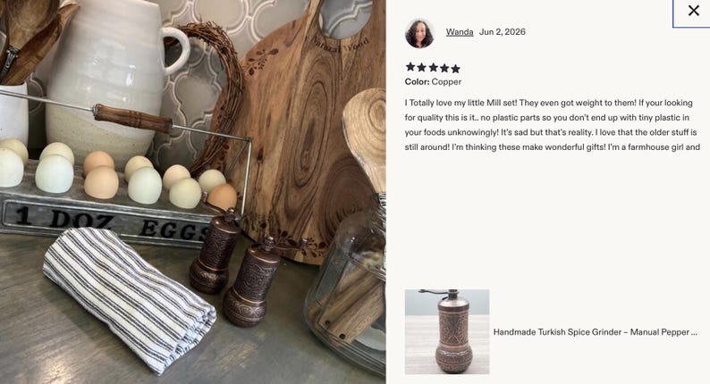 May include: Two cylindrical copper spice grinders with intricate embossed patterns, featuring a manual crank handle on top. The grinders are positioned next to a folded white and black striped cloth napkin, a metal egg crate labeled 1 DOZ EGGS, and a wooden board.