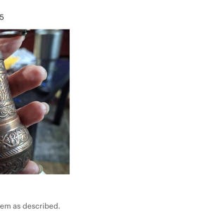 May include: A cylindrical metal spice grinder featuring an intricate embossed floral pattern across its surface. The grinder has a metallic finish with darkened recessed areas and a small crank handle attached to the top cap for manual operation.