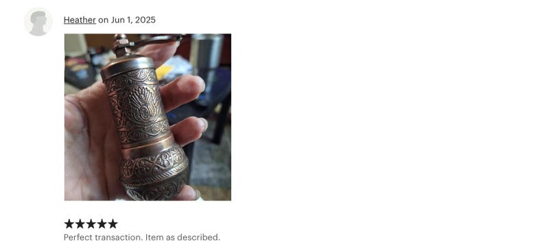 May include: A cylindrical metal spice grinder featuring an intricate embossed floral pattern across its surface. The grinder has a metallic finish with darkened recessed areas and a small crank handle attached to the top cap for manual operation.