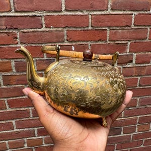 Puede incluir: Una tetera redonda de latón con un intrincado grabado floral en toda su superficie. La tetera incluye un pico curvo, un asa articulada envuelta en ratán de color claro y un pequeño pomo marrón oscuro en la tapa, apoyada sobre tres patas cortas.