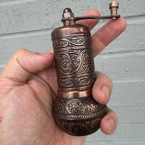 May include: A cylindrical copper-toned metal spice grinder featuring intricate embossed floral and scrollwork patterns throughout the body. The device includes a top-mounted manual crank handle with a small knob for operation and a rounded base.