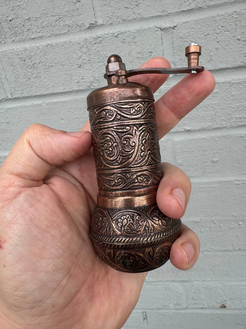 May include: A cylindrical copper-toned metal spice grinder featuring intricate embossed floral and scrollwork patterns throughout the body. The device includes a top-mounted manual crank handle with a small knob for operation and a rounded base.