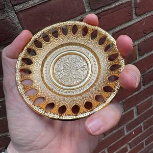 Puede incluir: Un platillo de metal dorado con un diseño decorativo y calado alrededor del borde. El platillo tiene un centro elevado con un patrón floral.