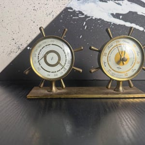 May include: A vintage brass ship wheel barometer and hygrometer set. The instruments have white faces with black markings and gold-colored hands. The set is mounted on a rectangular brass base.