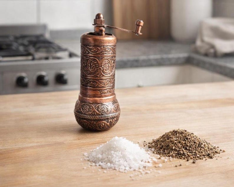 May include: A cylindrical copper spice grinder with an intricate embossed floral pattern and a top-mounted manual crank handle. Beside the grinder are two separate small piles of coarse white salt and ground black pepper on a wooden surface.