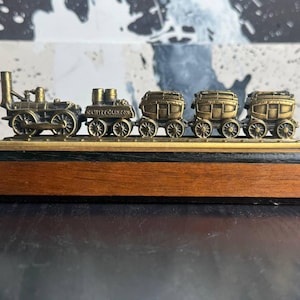May include: A brass-colored miniature train set, including a locomotive and several passenger cars, mounted on a wooden base. The train features detailed wheels and carriage designs, showcasing a vintage aesthetic. The base is rectangular with a dark top and a lighter wood finish.