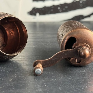 May include: A copper-colored metal spice grinder with an intricate embossed floral pattern. The device is shown in two parts: a hollow cylindrical base with an open top and a main body featuring a side-mounted hand crank with a small knob handle.