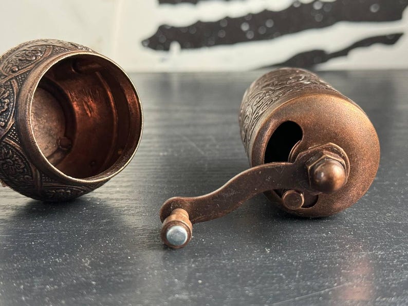 May include: A copper-colored metal spice grinder with an intricate embossed floral pattern. The device is shown in two parts: a hollow cylindrical base with an open top and a main body featuring a side-mounted hand crank with a small knob handle.
