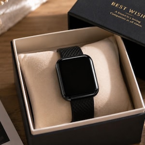 Peut inclure: Une montre connectée noire avec un bracelet en silicone texturé repose sur un coussin crème dans une boîte cadeau noire. Le couvercle porte le texte doré BEST WISHES et A friend is a loving Companion at all times, avec une doublure noire.