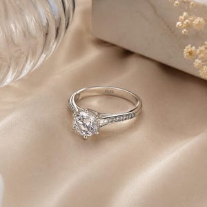 Peut inclure: Une bague argentée ornée d'une grande pierre centrale ronde et transparente à facettes, fixée par un sertissage à six griffes. L'anneau est agrémenté de petites pierres transparentes sur les côtés et porte l'inscription S925 à l'intérieur.