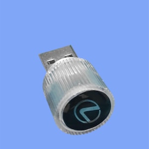 May include: A clear USB flash drive with a silver connector. The cylindrical body has a ridged texture and a black end cap with a blue logo. The background is a solid light blue.