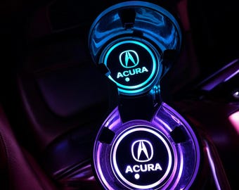 May include: Two circular cup holders integrated into a vehicle console, each featuring a glowing Acura logo. The top cup holder emits a bright cyan light, while the bottom cup holder glows in a vibrant purple hue, illuminating the interior space.