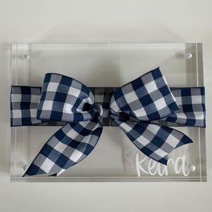 May include: A rectangular clear acrylic plaque featuring a navy blue and white gingham ribbon tied horizontally across the center. The name Keira is printed in white script lettering on the bottom right corner of the acrylic surface.