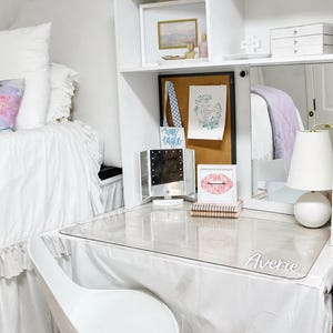 May include: A white desk with a clear top and the name "Averie" in script. A white chair with gold legs sits in front of the desk. A lighted makeup mirror, a notebook, and a framed print are on the desk. A bulletin board and a lamp are also visible.