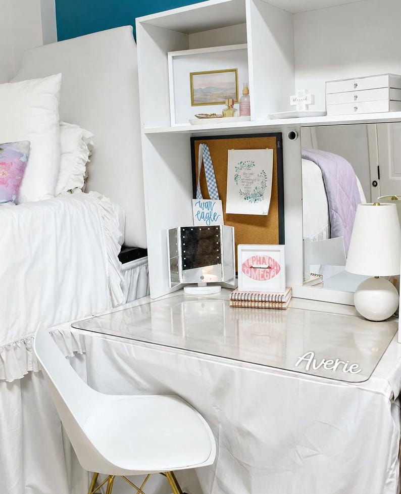 May include: A white desk hutch with a clear acrylic desk mat featuring the name Averie. The setup includes a cork board, a tri-fold vanity mirror, a white lamp, a small framed print with the text Alpha Chi Omega, and a white chair.