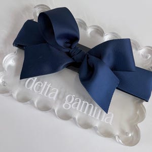 May include: A dark navy blue grosgrain ribbon bow attached to a clear, scalloped-edge acrylic rectangular plaque. The plaque features the white text delta gamma printed in a lowercase serif font across the center of the transparent surface.