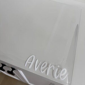 May include: A clear, rectangular acrylic sheet featuring the name Averie in white, cursive-style lettering. The transparent material is positioned on a white surface, showcasing the custom white vinyl decal applied to the smooth, flat surface.