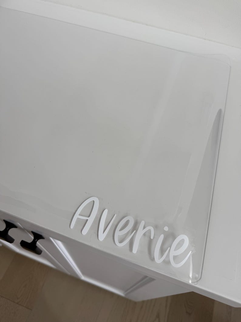 May include: A clear, rectangular acrylic sheet featuring the name Averie in white, cursive-style lettering. The transparent material is positioned on a white surface, showcasing the custom white vinyl decal applied to the smooth, flat surface.