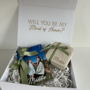 May include: A white gift box with the text WILL YOU BE MY Maid of Honor? printed in gold inside the lid. Inside are a photo frame featuring two people and the name Maddie, and a beige square jewelry case with a green ribbon and the name Maddie.