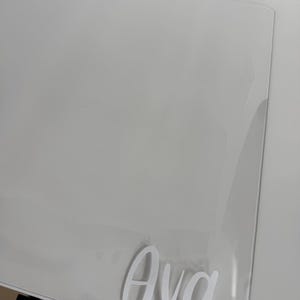 May include: A clear, rectangular acrylic sheet featuring the name Ava in white, cursive-style lettering applied to the surface. The transparent material has smooth, rounded corners and a glossy finish, reflecting light across its flat, clean surface.